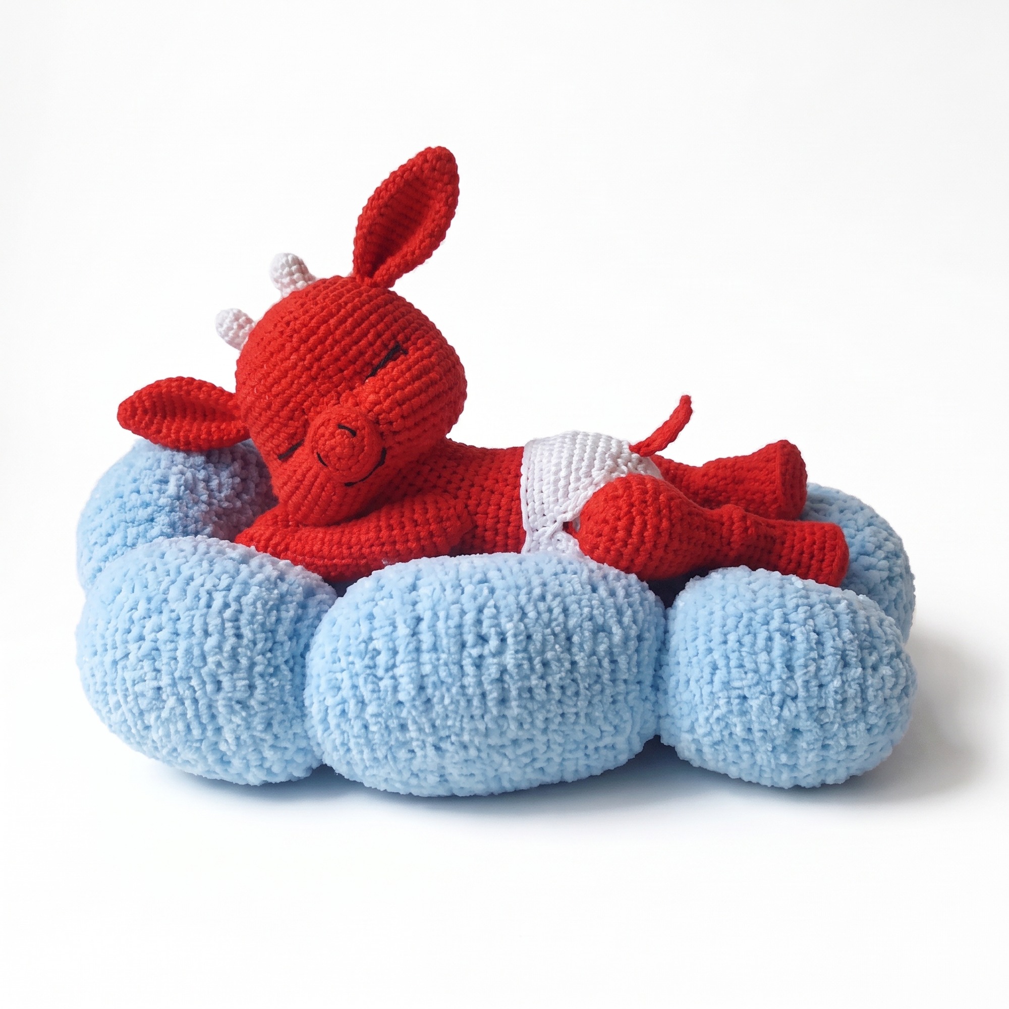 The finished crochet devil pattern — a red amigurumi devil in a white nappy lying on a soft blue knitted cloud