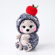 The finished crochet hedgehog pattern — a grey fluffy amigurumi hedgehog with a cream velvet face and a red crocheted apple balanced on his head